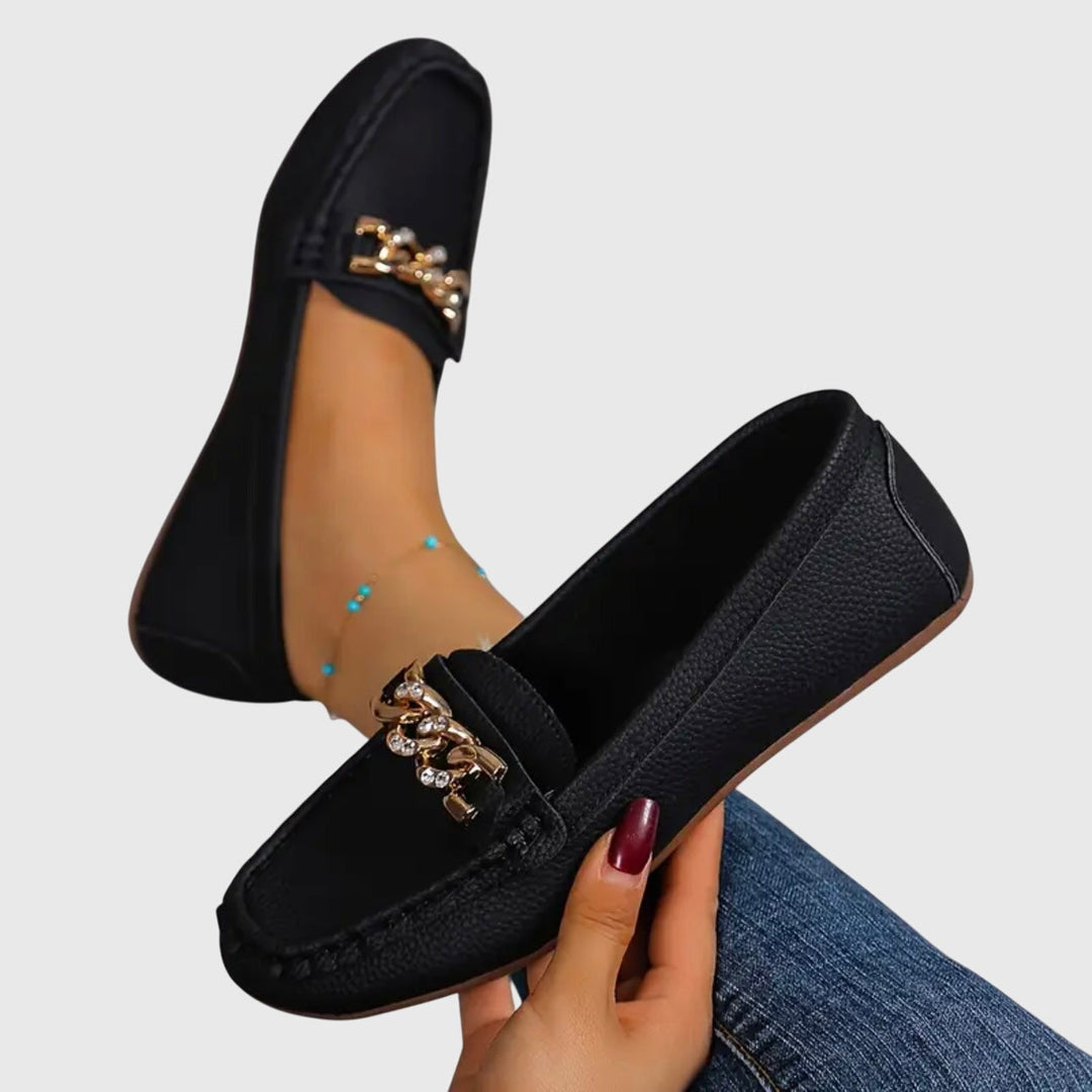 Paar zwarte dames loafers met gouden ketting en platte zool. Modieuze en comfortabele instapschoenen.